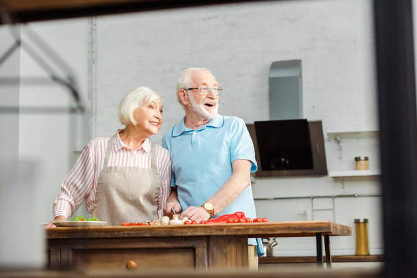 Selektywne skupienie się na uśmiechniętej starszej parze odwracającej wzrok podczas krojenia świeżych warzyw na stole kuchennym - Zdjęcie, obraz