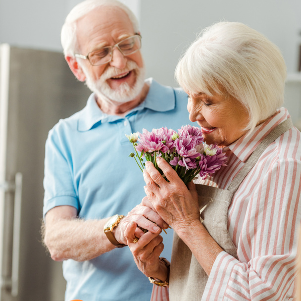 Selective focus of senior woman in apron smelling bouquet by smiling husband in kitchen  - Photo, Image