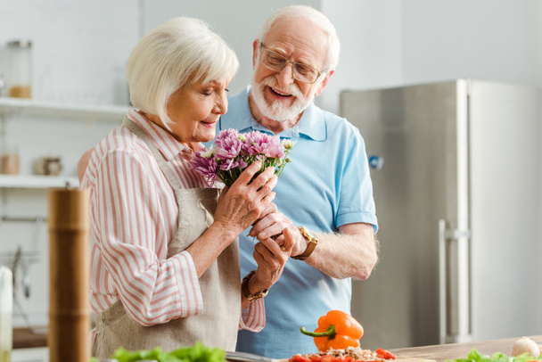 キッチンテーブルの上で笑顔の夫と野菜の横に花束を嗅ぐシニア女性の選択的な焦点  - 写真・画像