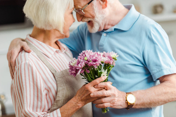 Selective focus of senior man hugging smiling wife with bouquet of chrysanthemums - Fotó, kép