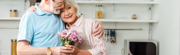 Panoramic shot of senior man hugging smiling wife with bouquet of chrysanthemums in kitchen  - Photo, Image