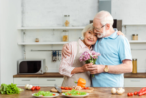 Älterer Mann umarmt lächelnde Frau mit Strauß neben reifem Gemüse auf Küchentisch - Foto, Bild