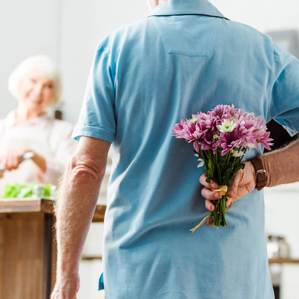 Selektivní zaměření seniora muž skrývá kytice, zatímco se usmívá manželka vaření v pozadí - Fotografie, Obrázek