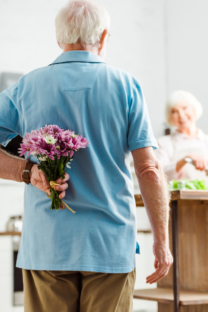 Selective focus of senior man hiding floral bouquet while smiling wife cooking in kitchen - Photo, Image