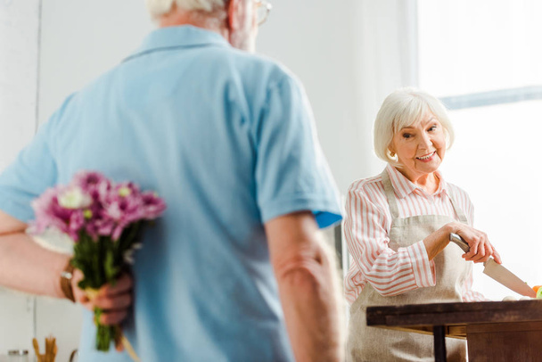 Selectieve focus van lachende oudere vrouw op zoek naar echtgenoot met boeket tijdens het koken op keukentafel  - Foto, afbeelding