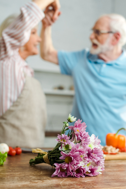 Selective focus of bouquet and vegetables on kitchen table and senior couple dancing at background - 写真・画像