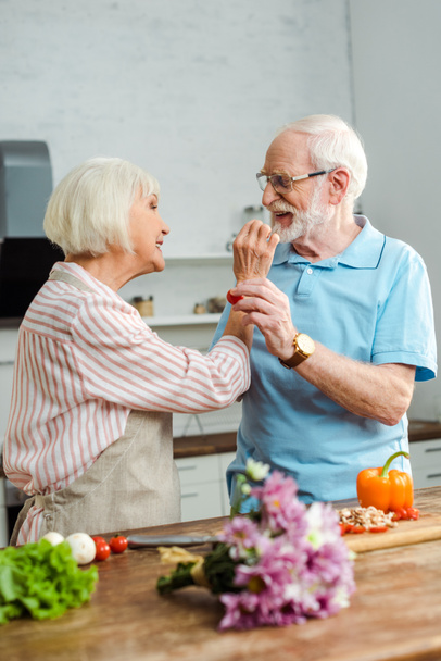 Az egymást meggyparadicsommal etető idős házaspár szelektív fókusza zöldségekkel és csokorral a konyhaasztalon - Fotó, kép