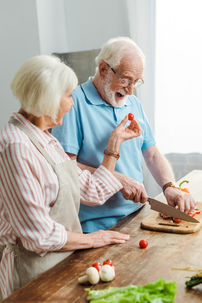 Side näkymä vanhempi nainen antaa kirsikka tomaatti aviomies samalla leikkaamalla vihanneksia keittiön pöydälle
 - Valokuva, kuva