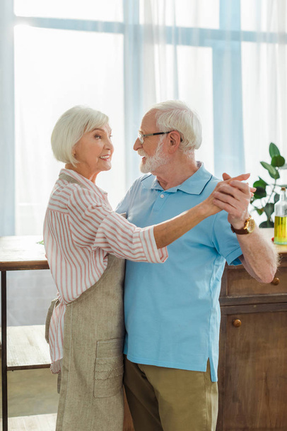Вид сбоку на пожилого человека, танцующего с улыбающейся женой в фартуке на кухне
 - Фото, изображение