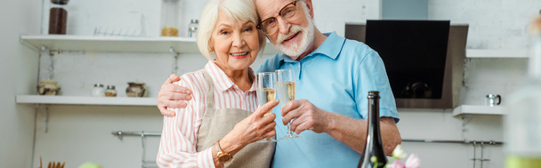 Πανοραμική φωτογραφία του ηλικιωμένου ζευγαριού να ψήνει με σαμπάνια και να κοιτάζει την κάμερα στην κουζίνα - Φωτογραφία, εικόνα