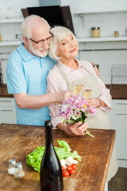 Selective focus of elderly woman holding bouquet and toasting champagne with husband in kitchen  - Photo, Image