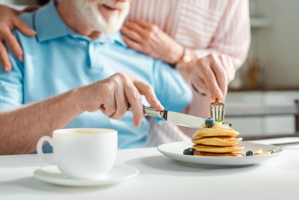 Cropped view of senior man cutting pancakes beside wife in kitchen - 写真・画像