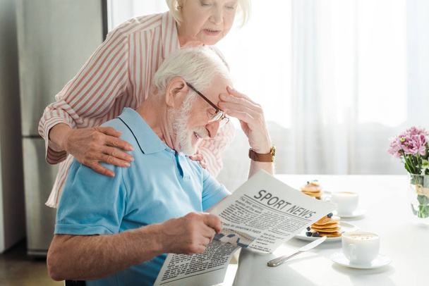 Oldalnézet idős férfi olvas újságot sport hírek feleség reggeli közben - Fotó, kép