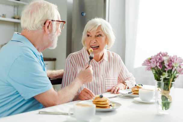 Επιλεκτική εστίαση του ηλικιωμένου άνδρα σίτιση γυναίκα με τηγανίτα κατά τη διάρκεια του πρωινού στην κουζίνα - Φωτογραφία, εικόνα