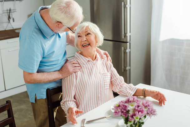 Valikoiva painopiste vanhempi mies syleilee hymyilevä vaimo pöydässä keittiössä
 - Valokuva, kuva