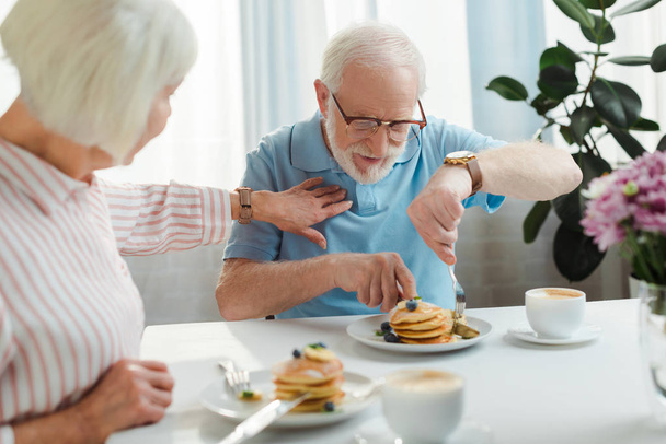 Enfoque selectivo de la mujer mayor tocando marido con panqueques por café y flores en la mesa
 - Foto, imagen