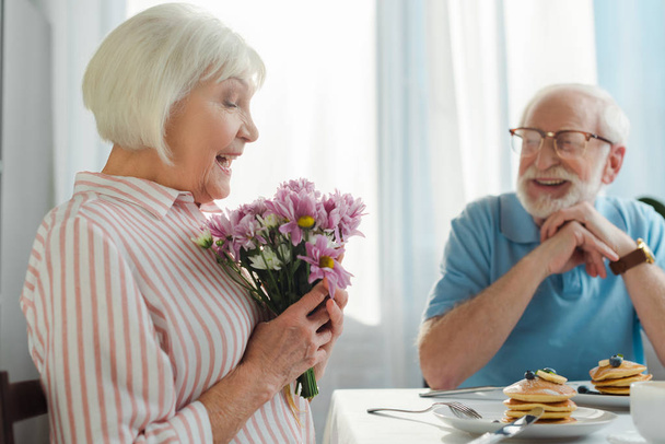 Selective focus of woman holding bouquet by smiling husband at table with coffee and pancakes - Foto, Imagem