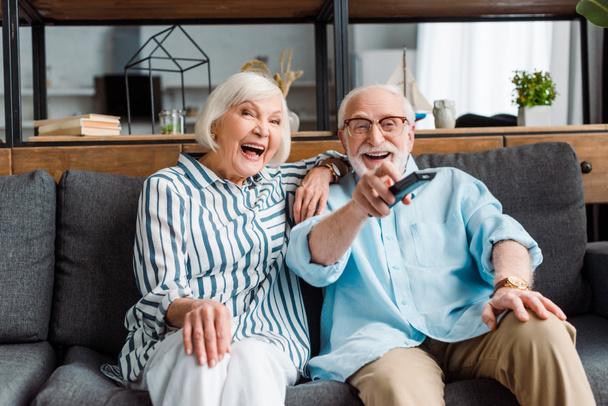 Senior couple laughing while watching tv on couch in living room - Foto, Imagen