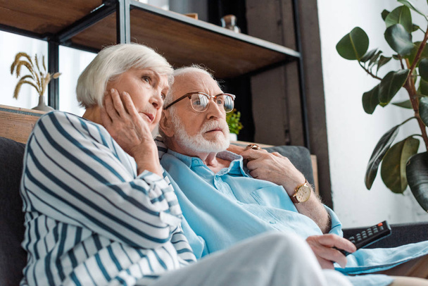 Low angle view of serious senior couple watching tv on sofa at home - Valokuva, kuva