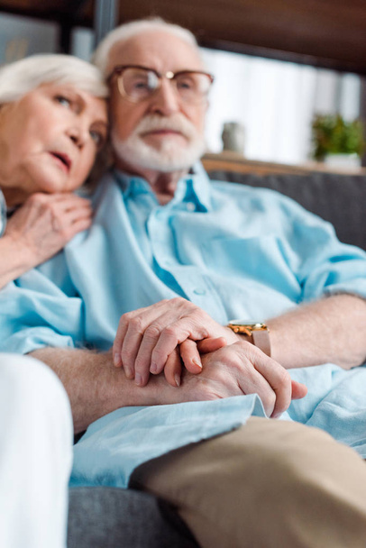 Selective focus of senior couple holding hands while sitting on sofa at home - 写真・画像