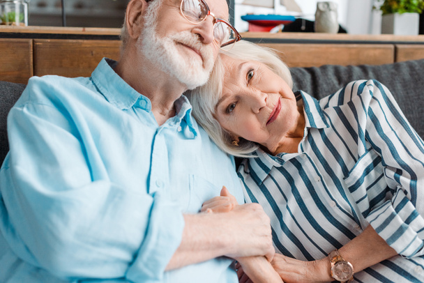 Senior woman looking at camera while holding hand of husband on sofa - Фото, изображение