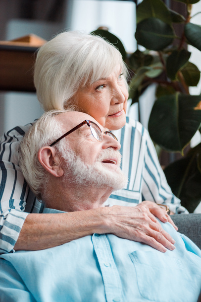 Senior woman hugging husband and looking away at home - Foto, afbeelding
