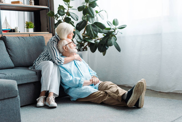 Senior woman embracing husband on floor and looking away in living room - Фото, зображення