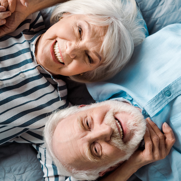 Vista superior de la pareja de ancianos sonriendo a la cámara mientras están acostados en la cama
 - Foto, Imagen