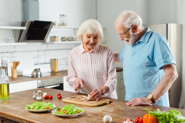 Літній чоловік обіймає усміхнену дружину, що ріже гриби за кухонним столом
 - Фото, зображення
