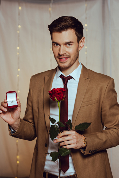 elegant, handsome man holding box with wedding ring and red rose while smiling at camera in restaurant - Фото, зображення