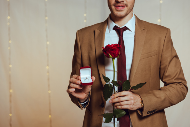 partial view of elegant man holding box with wedding ring and red rose while standing in restaurant - Фото, изображение