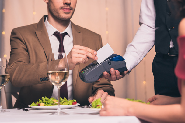vista ritagliata di uomo elegante che paga con carta di credito con terminale mentre è seduto in ristorante vicino alla fidanzata
 - Foto, immagini