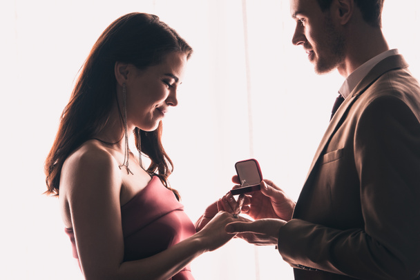 zijaanzicht van knappe man presenteren ring naar gelukkig vriendin terwijl het maken van huwelijksaanzoek op valentijnsdag - Foto, afbeelding