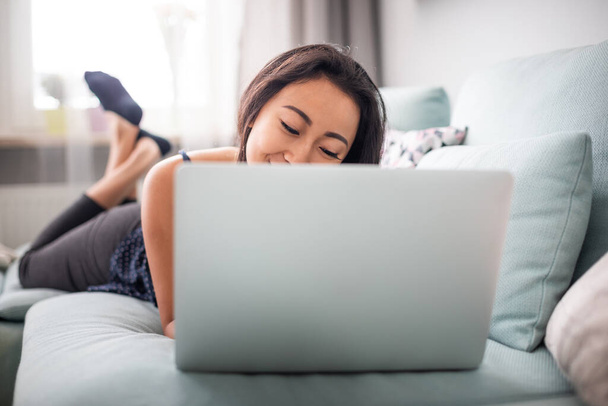 Smiling asian young woman using laptop lying on sofa at home - Foto, Imagem
