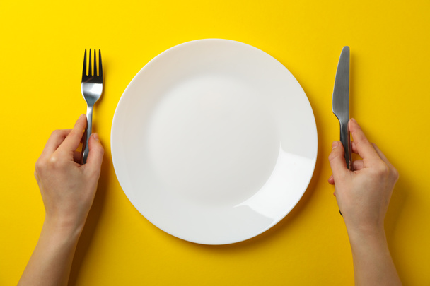 Vrouwelijke handen houden vork en mes op gele achtergrond met plaat - Foto, afbeelding