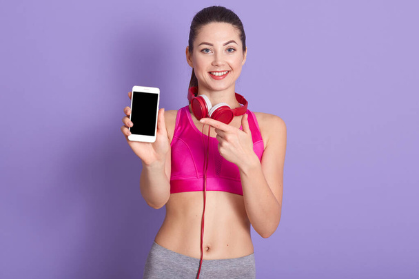 Image of cheerful sporty adorable female making gesture, pointing at smartphone with blank screen, holding her device in one hand, smiling sincerely, being in high spirits. Technology concept. - Photo, image