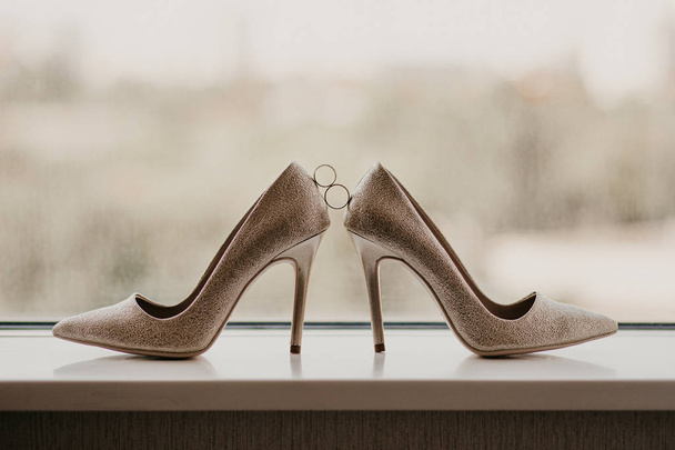 Χρυσές βέρες ανάμεσα σε χρυσά ψηλοτάκουνα παπούτσια. Λεπτομέρειες γάμου στον καθρέπτη κοντά σε πανοραμικό παράθυρο - Φωτογραφία, εικόνα