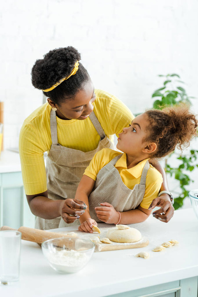 Χαρούμενη Αφροαμερικανή μητέρα που κοιτάζει αξιολάτρευτο παιδί και σμιλεύει ζυμαρικά στην κουζίνα  - Φωτογραφία, εικόνα
