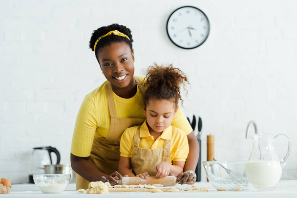 cute african american kid holding rolling pin near cheerful mother and dumplings on cutting board  - Foto, Imagen