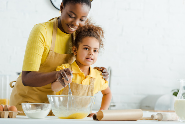 かわいいアフリカ系アメリカ人の子供と陽気な母親が台所で料理をしながらボウルの近くに泡立て器を保持  - 写真・画像