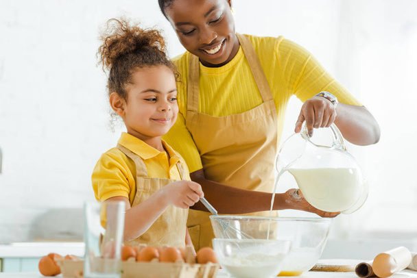 selective focus of happy african american mother pouring milk in bowl near cute daughter  - Foto, afbeelding