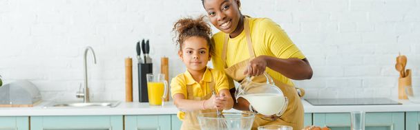 panoramic shot of cheerful african american mother pouring milk in bowl near cute daughter and looking at camera - Photo, Image