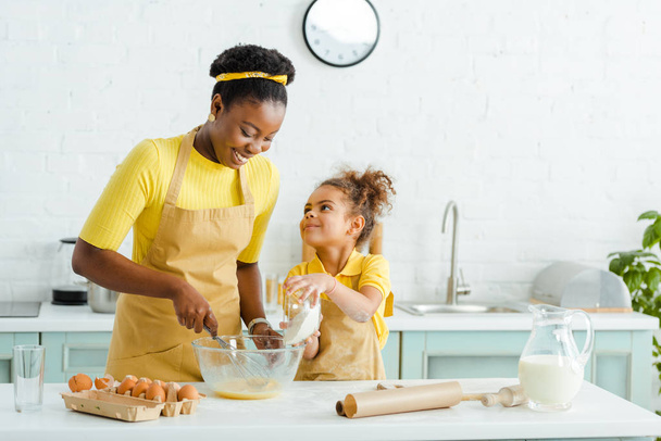 süßes afrikanisches amerikanisches Kind schaut Mutter an, während es Schüssel mit Mehl hält  - Foto, Bild