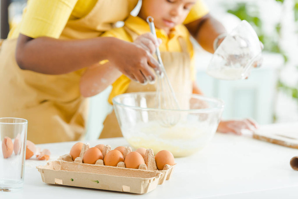 selectieve focus van eieren in de buurt van Afro-Amerikaanse kind en moeder mengen ingrediënten in kom  - Foto, afbeelding
