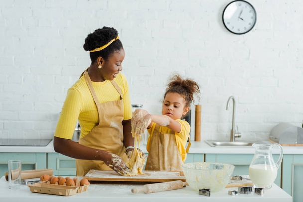 χαρούμενη αφρικάνικη μητέρα και κόρη ζυμώνουν ζύμη κοντά στα υλικά της κουζίνας  - Φωτογραφία, εικόνα