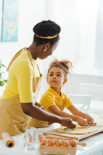 mignon afro-américain enfant regardant mère tout en tenant des emporte-pièces près de pâte crue dans la cuisine
  - Photo, image
