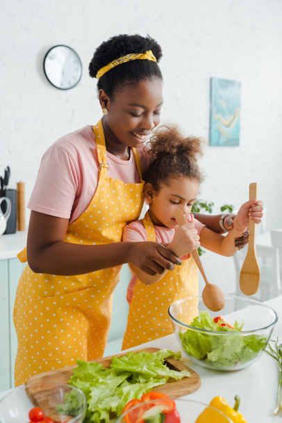 cheerful african american mother and cute child holding wooden spoon and spatula while preparing salad  - Foto, imagen