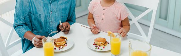 panoramic shot of african american mother and daughter holding cutlery near pancakes  - Zdjęcie, obraz