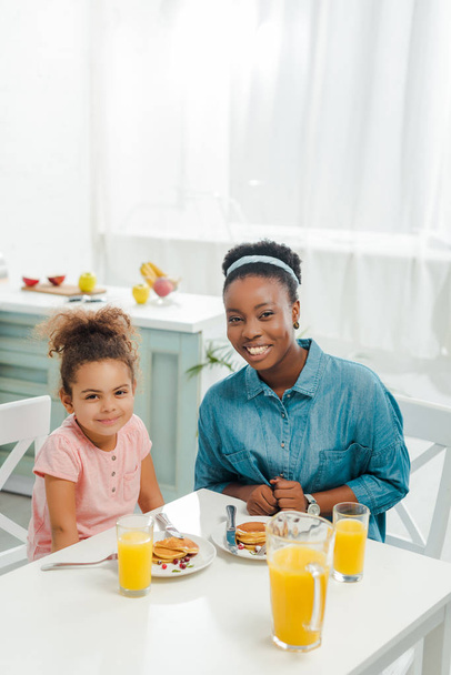 cheerful african american mother and daughter looking at camera near tasty pancakes  - Fotografie, Obrázek