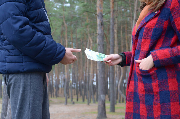Una chica transfiere billetes de euro a manos de un joven en el bosque. Concepto de robo o transacción ilegal
 - Foto, imagen